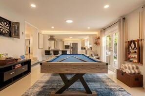 Een pooltafel in een moderne ruimte, met een keuken op de achtergrond, een dartbord en een plank met ballen.