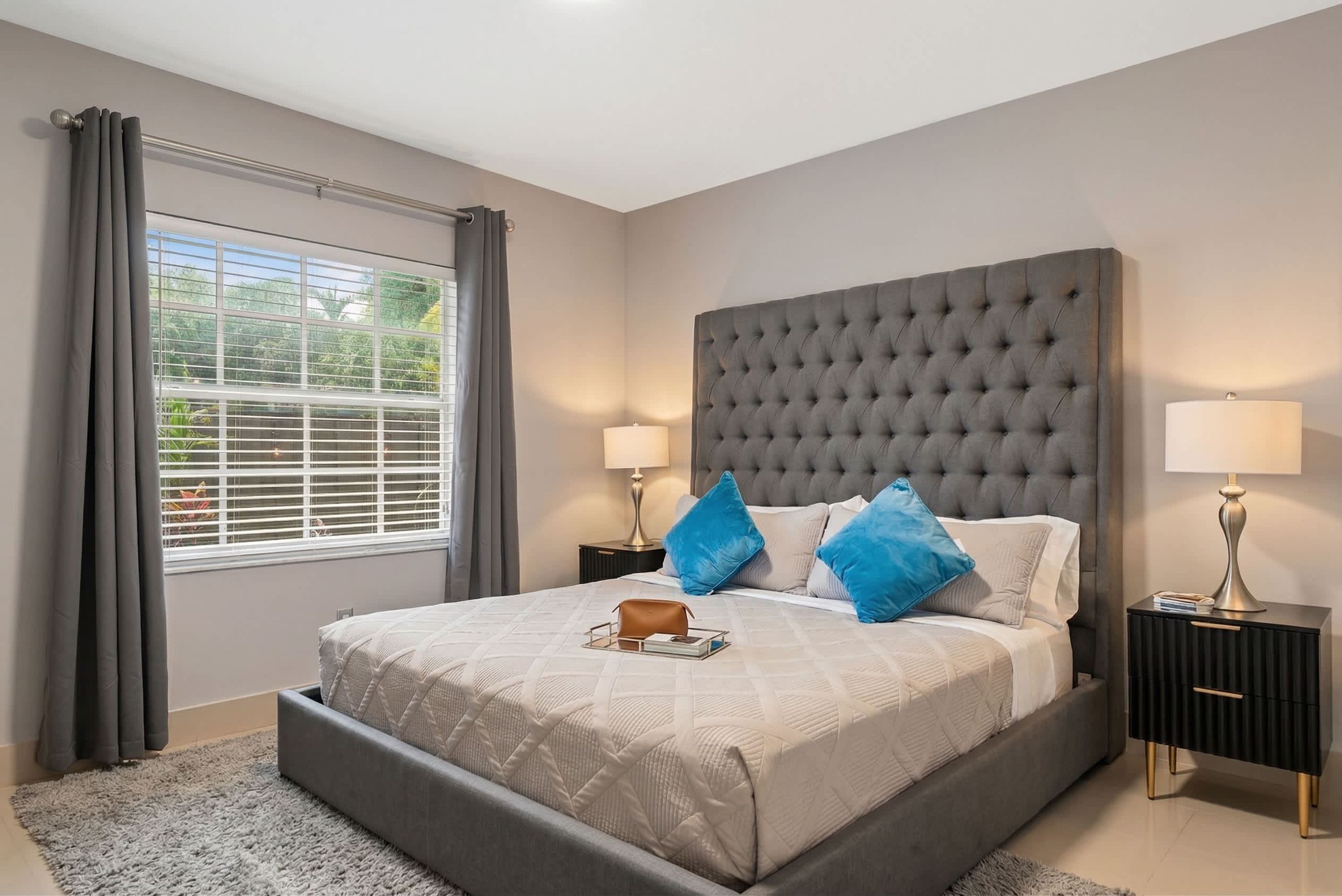 A bedroom with a large bed, two bedside tables with lamps, a window with curtains, and a view of greenery outside.