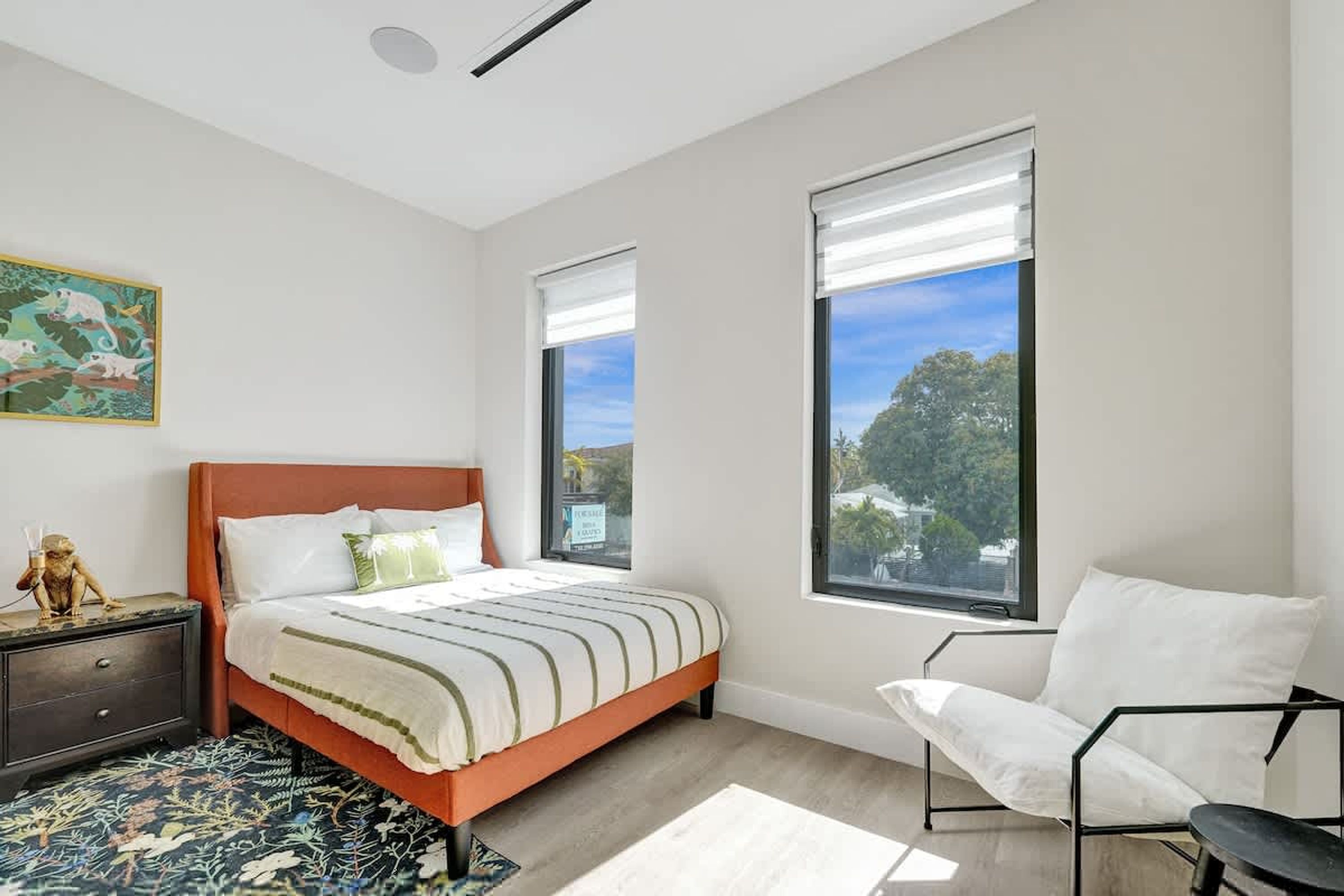 A bedroom with a bed, a chair, a nightstand, a painting, and large windows.