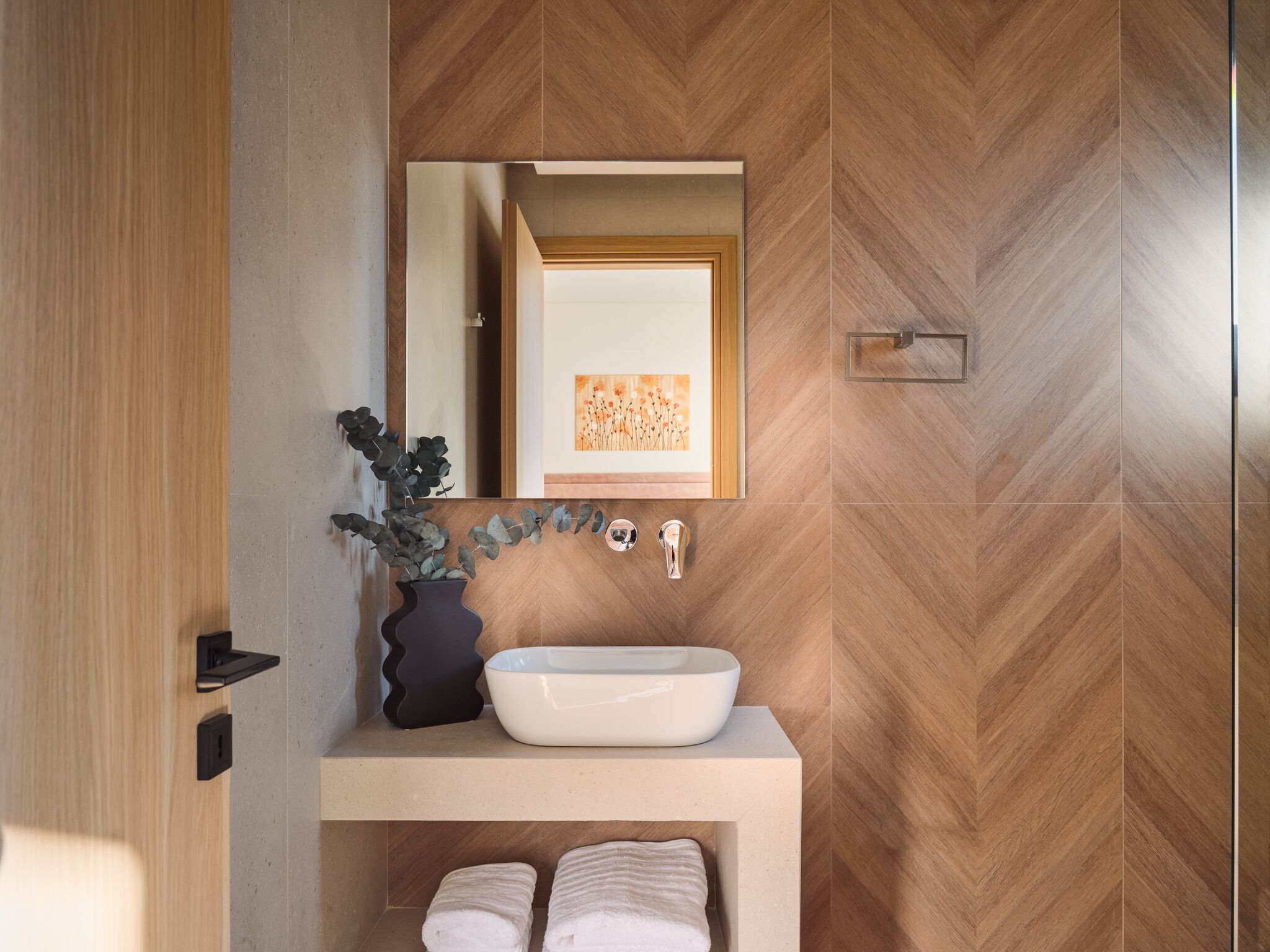 Une salle de bain avec un mur en bois disposé en motif chevron, un lavabo sur colonne blanc, un vase noir avec une plante, un miroir et un porte-serviettes.