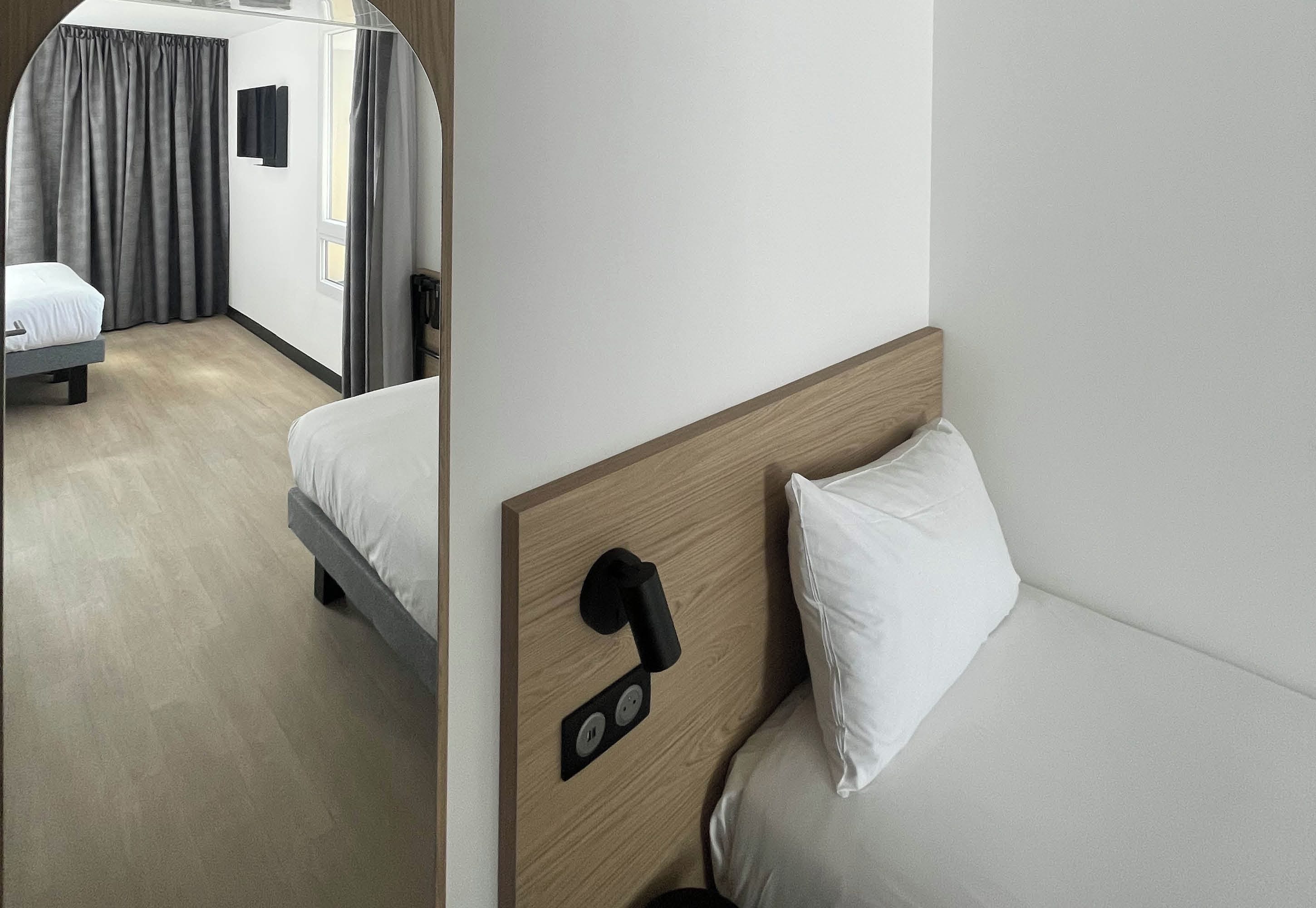 Une chambre d’hôtel avec un lit, une table de chevet, une prise de courant fixée au mur et une fenêtre avec des rideaux.