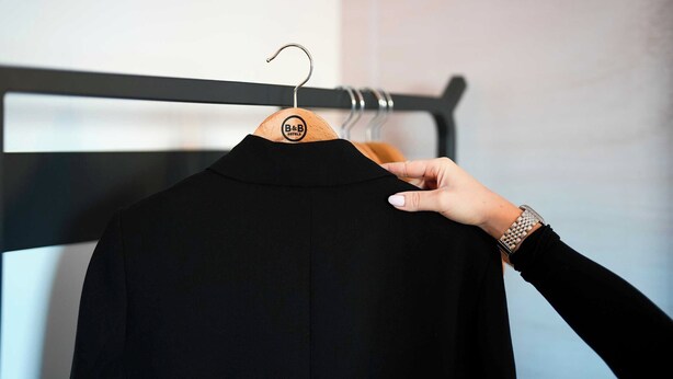 A black garment on a wooden hanger with a logo, held by a hand with a watch.
