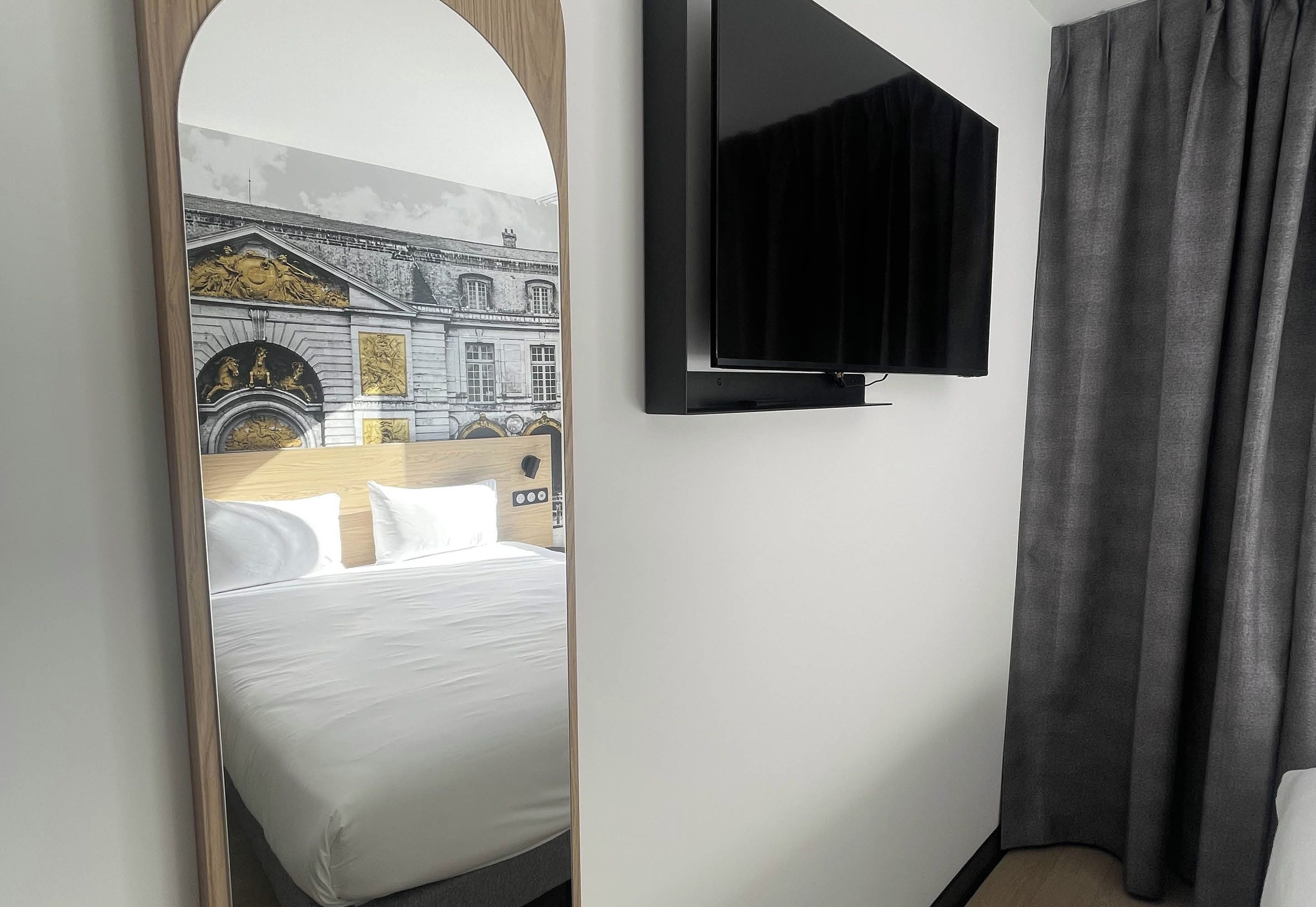 Ein Hotelzimmer mit einem Bett, einem an der Wand befestigten Fernseher und einem großen Spiegel, der eine Gebäudefassade reflektiert.