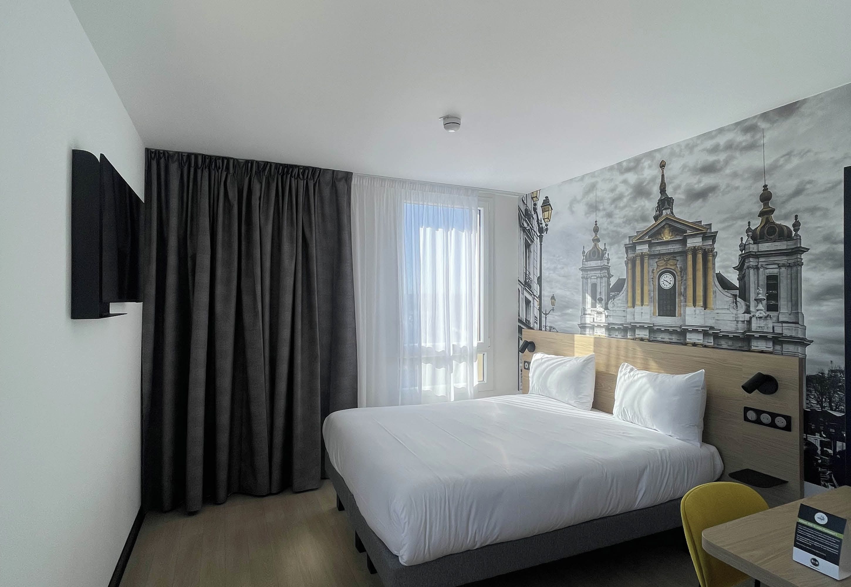 Une chambre d’hôtel avec un lit, un téléviseur fixé au mur, une fenêtre avec des rideaux et une fresque représentant un bâtiment.