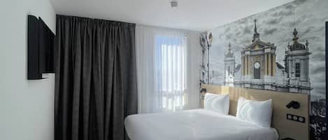 Habitación de hotel con cama, televisión instalada en la pared, ventana con cortinas y un mural de un edificio.