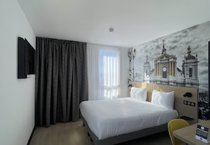 Habitación de hotel con cama, televisión instalada en la pared, ventana con cortinas y un mural de un edificio.