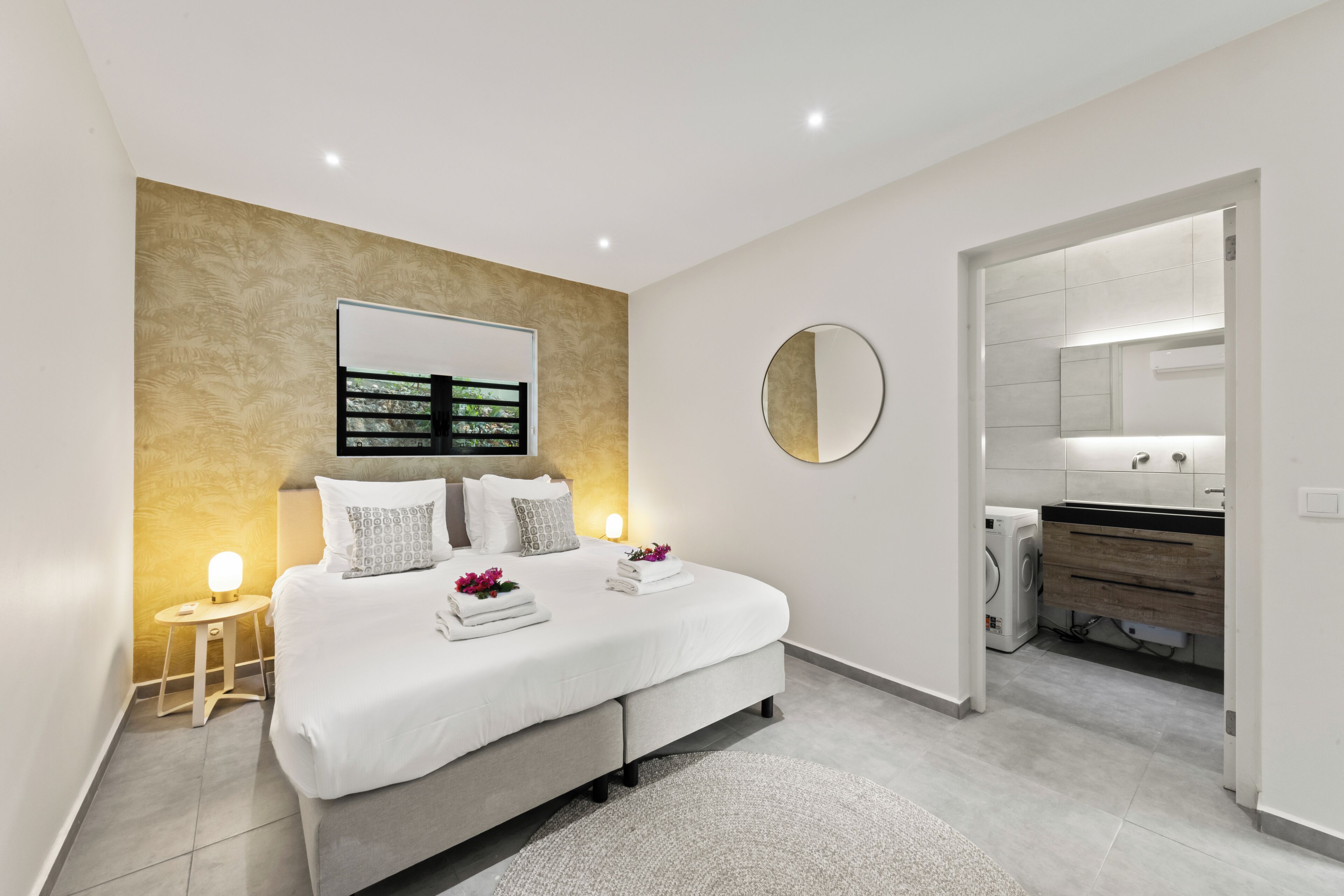 A modern bedroom with a bed, bedside tables, a wall-mounted TV, and a bathroom visible through an open door.