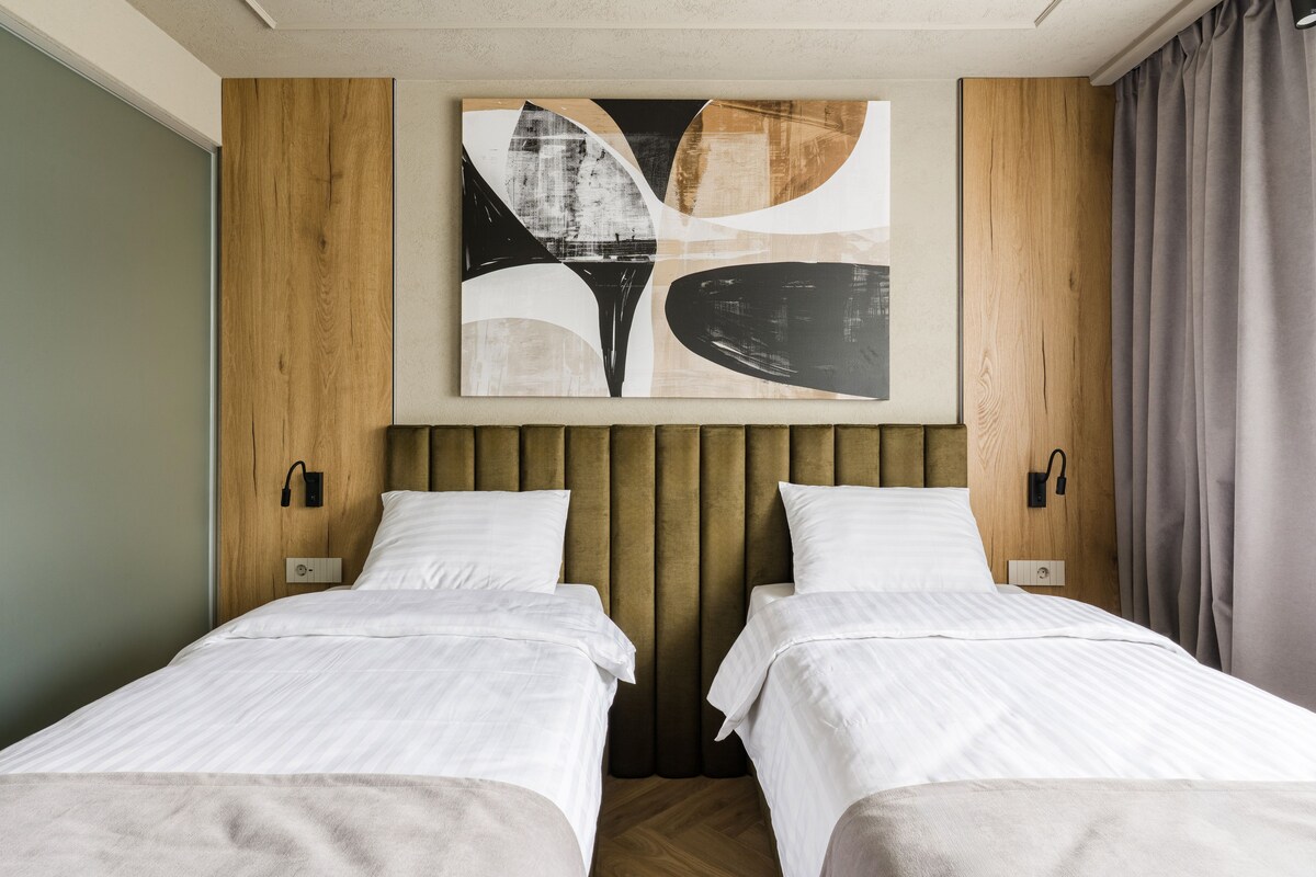 Habitación de hotel con dos camas, un cuadro abstracto moderno, paneles de madera y vistas a la ciudad.