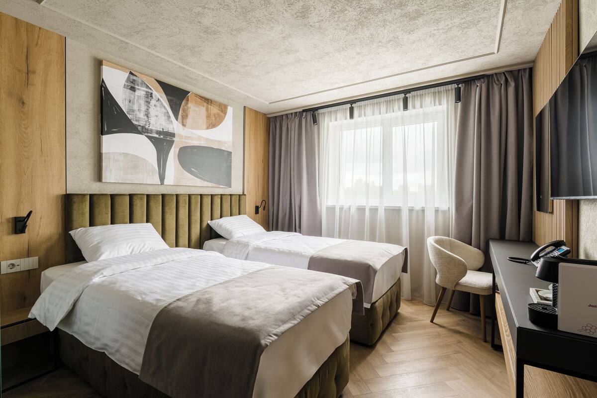 A hotel room with two beds, a desk with a laptop, a chair, and a large abstract painting on the wall.