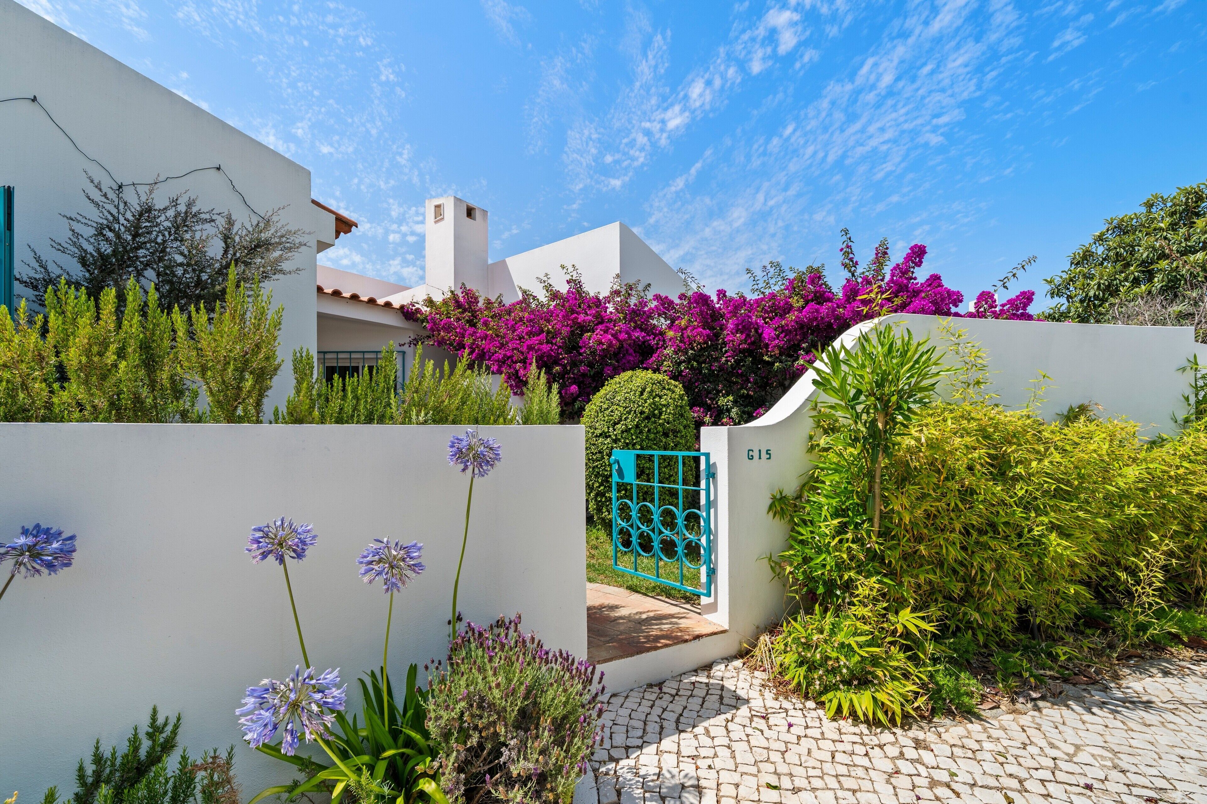 A modern house with a white exterior, a blue gate, and lush greenery including purple bougainvillea.
