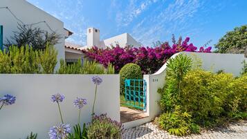 A modern house with a white exterior, a blue gate, and lush greenery including purple bougainvillea.