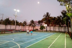 Een buitenbasketbalveld met een basketbalring, twee spelers en palmbomen op de achtergrond.