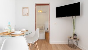 Un'area pranzo moderna con tavolo in vetro, sedie bianche, una TV a parete e un piccolo tavolino con una pianta.