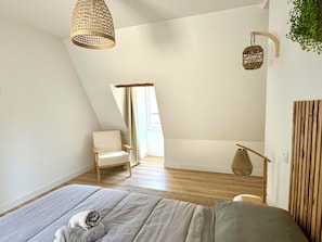 A bedroom with a sloped ceiling, a bed with a grey blanket, a chair, a hanging basket, and a wooden wall panel. - Caporizon—Les Balancoires—2 bedrooms—Right in the center (Sarlat-la-Canéda)