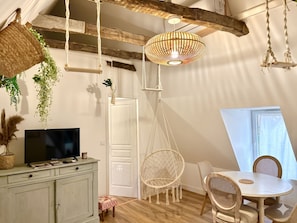 A rustic room with a hanging light fixture, a television on a cabinet, a dining table with chairs, and a door. - Caporizon—Les Balancoires—2 bedrooms—Right in the center (Sarlat-la-Canéda)
