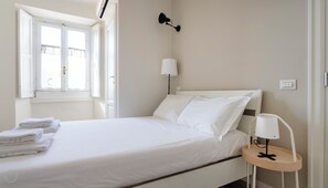 Ein ordentlich bezogenes Bett mit weißen Leinen, ein Nachttisch mit Lampe und ein Fenster mit Vorhängen.