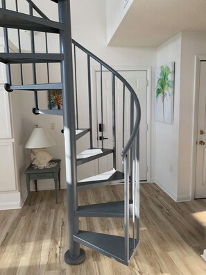 Eine Wendeltreppe mit Metallgeländer, eine weiße Tür im Hintergrund, ein kleiner Tisch mit einer Lampe und eine Wand mit einem Gemälde.