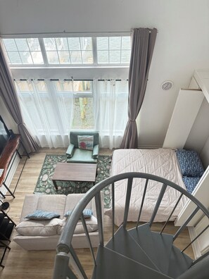 Ein Zimmer mit einem Bett, einem Schlafsofa, einem kleinen Tisch und einem Sessel. Ein Fenster mit Vorhängen, eine Uhr an der Wand und eine Heizung sind ebenfalls vorhanden.