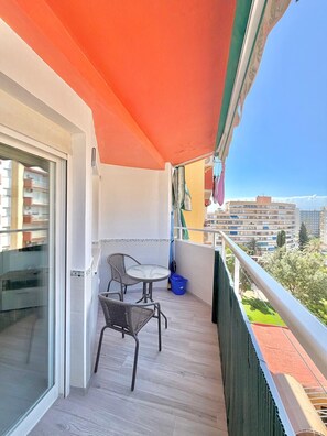 A balcony with a table and chairs, a blue trash bin, and a view of buildings and trees. - TERRASOL TRINIDAD APARTMENT (Torre del Mar)