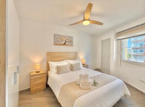A bedroom with a bed, wooden headboard, bedside lamps, a ceiling fan, a window with a view, and a welcome sign. - TERRASOL TRINIDAD APARTMENT (Torre del Mar)