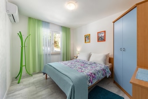 Ein Schlafzimmer mit Bett, Kleiderschrank, Standventilator und einem Fenster mit Vorhängen.