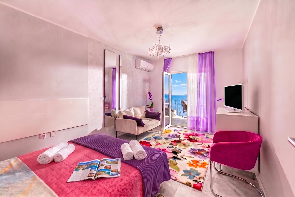A hotel room with a bed, a chair, a TV, and a view of the sea.