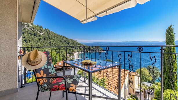 Uma varanda com mesa e cadeiras, um chapéu e vista para o mar e colinas.