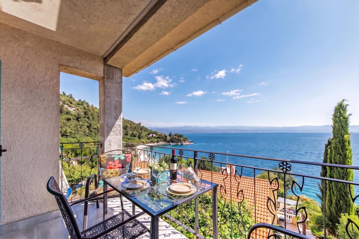 A balcony with a table set for dining, overlooking a coastal landscape with a clear sky.