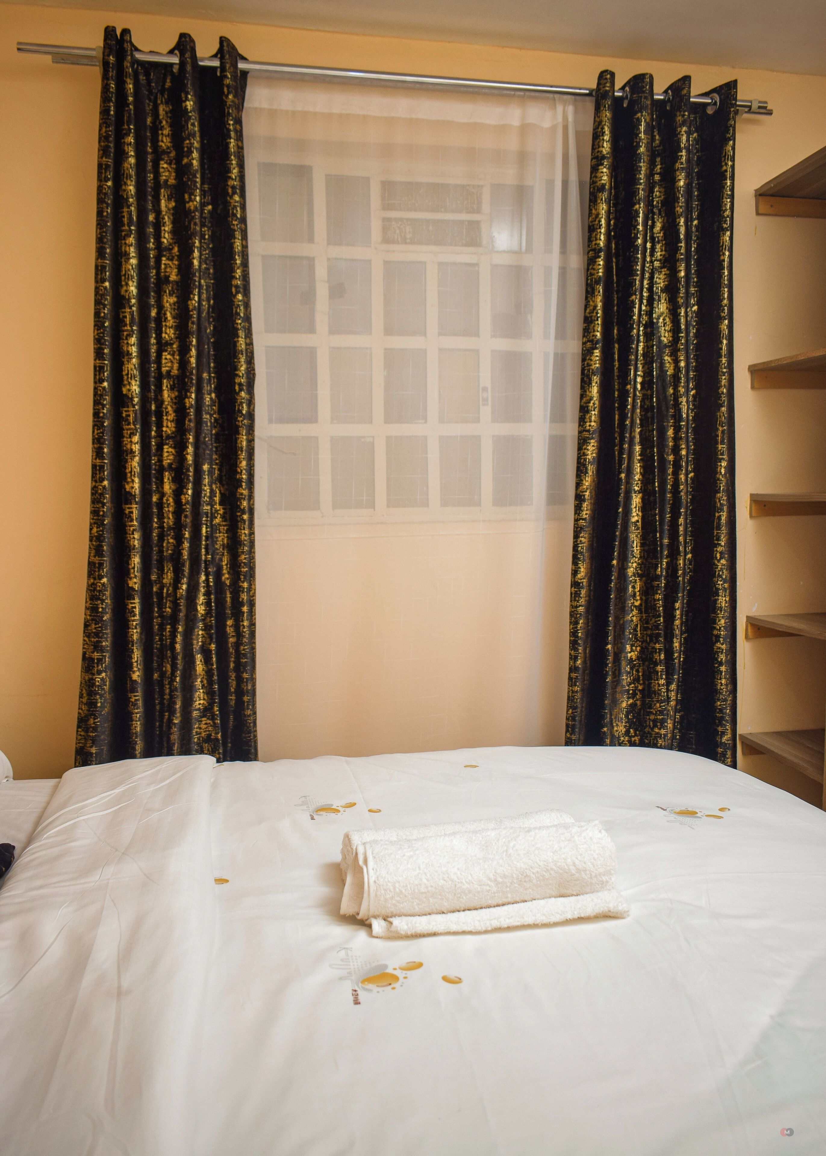 Een hotelkamer met een bed, wit beddengoed, een opgerolde witte handdoek en zwarte gordijnen met gouden patronen.