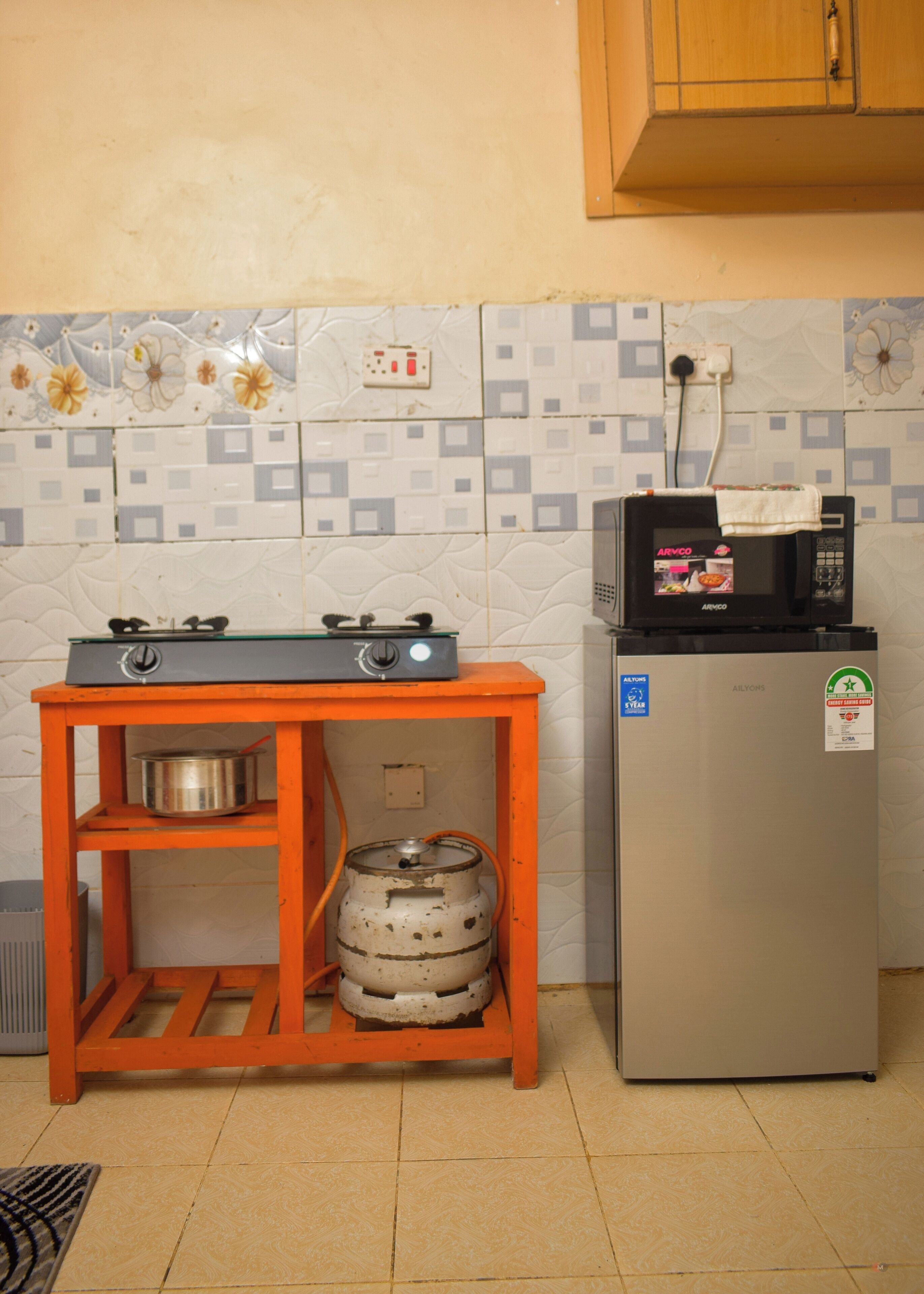 A kitchen with a gas stove, a microwave, a refrigerator, and a gas cylinder on a shelf.