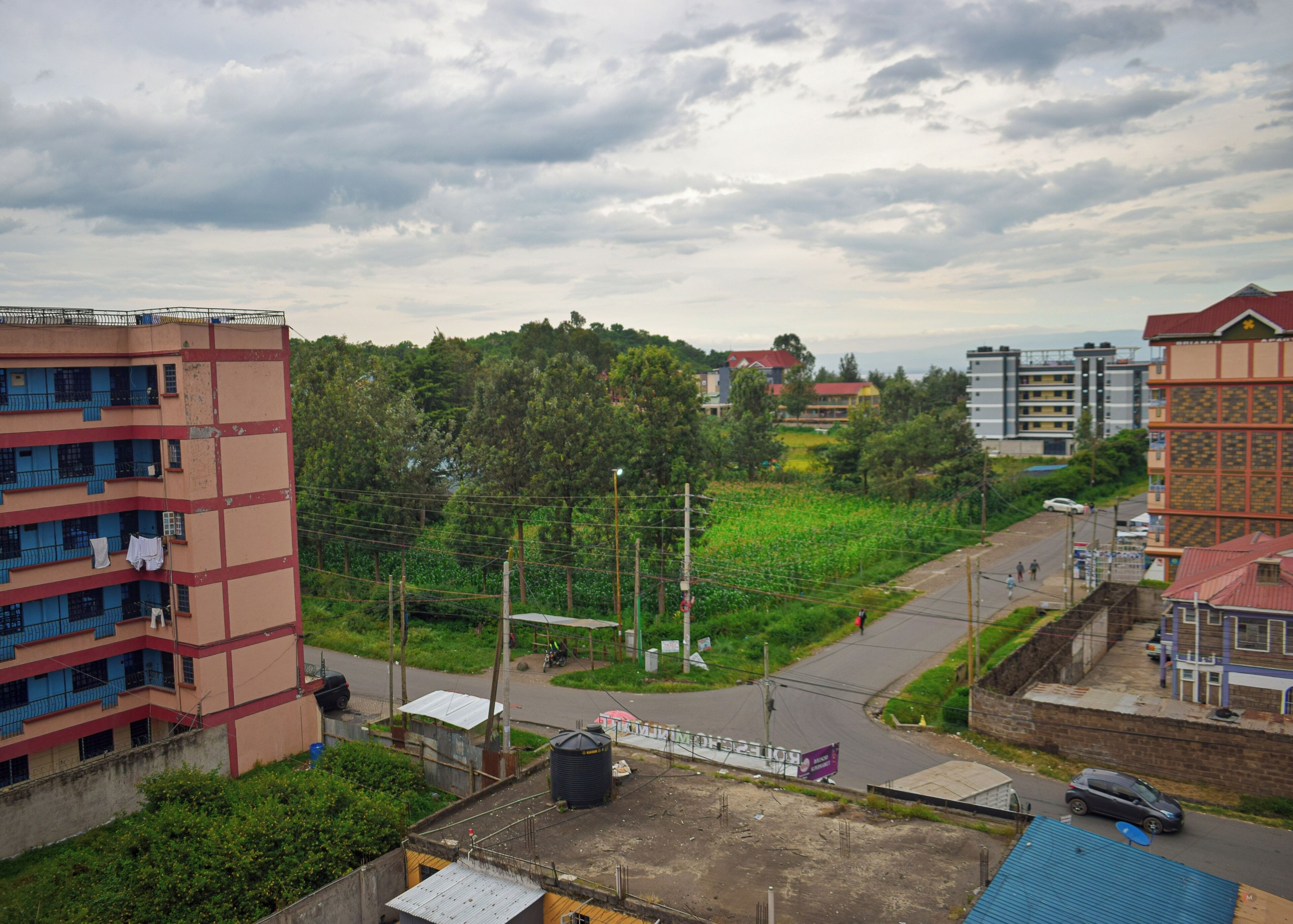 A multi-story building with balconies, a road, and a green area with trees.