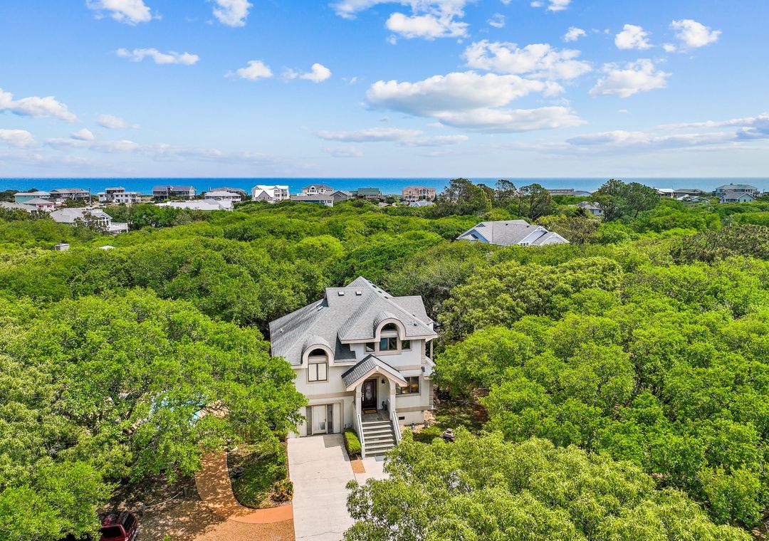 A large house surrounded by dense greenery, with a clear view of the ocean in the background.