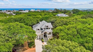 A large house surrounded by dense greenery, with a clear view of the ocean in the background. - 🌊 OBX Escape w/ Pool, Hot Tub & Volleyball Court — Pet Friendly 🐶 (Southern Shores)