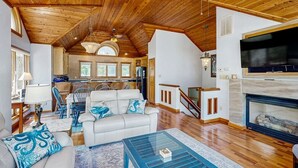 A spacious living area with a high ceiling, a fireplace, a television, and a kitchen in the background. - 🌊 OBX Escape w/ Pool, Hot Tub & Volleyball Court — Pet Friendly 🐶 (Southern Shores)