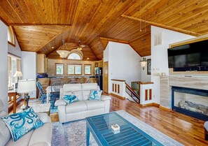 Ein geräumiger Wohnbereich mit hoher Decke, Kamin, Fernseher und einer Küche im Hintergrund.