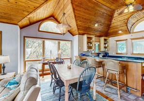 Ein gemütliches Interieur mit Holzbalkendecke, einer Essecke mit Stühlen und Tisch, einer Küche mit Bar und großen Fenstern.