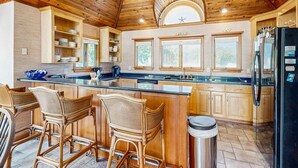 A kitchen with wooden cabinets, a central island with bar stools, a black refrigerator, and a large window with a view of trees. - 🌊 OBX Escape w/ Pool, Hot Tub & Volleyball Court — Pet Friendly 🐶 (Southern Shores)