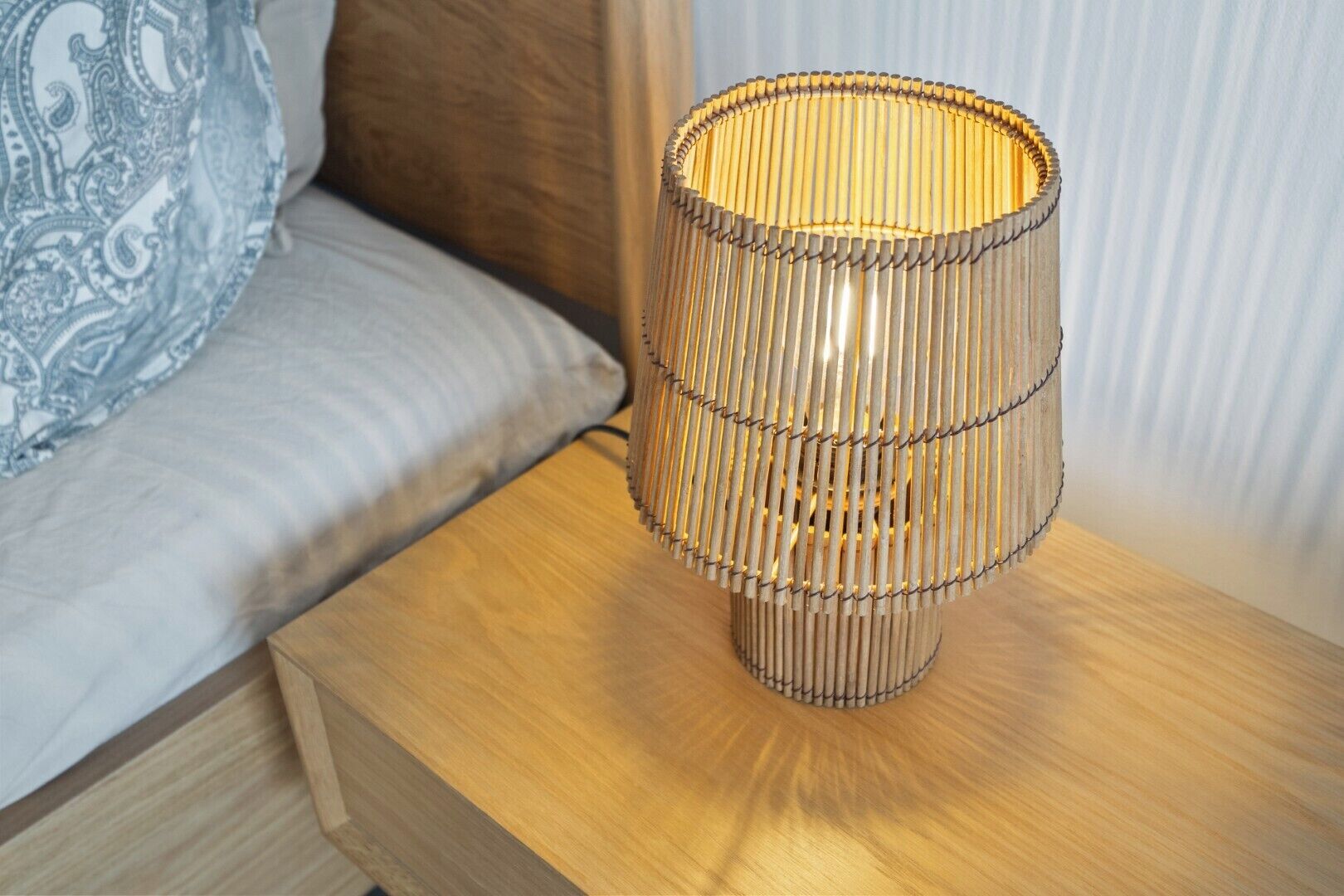 Une lampe de chevet au motif tissé, posée sur une table de chevet en bois, à côté d’un lit agrémenté d’un coussin à motifs.