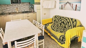 Eine Küche mit türkis-blauen Schränken, einer Mikrowelle und einer gefliesten Spritzschutzwand. Es gibt einen Esstisch mit Stühlen sowie ein gemustertes Sofa mit gelber Tagesdecke.