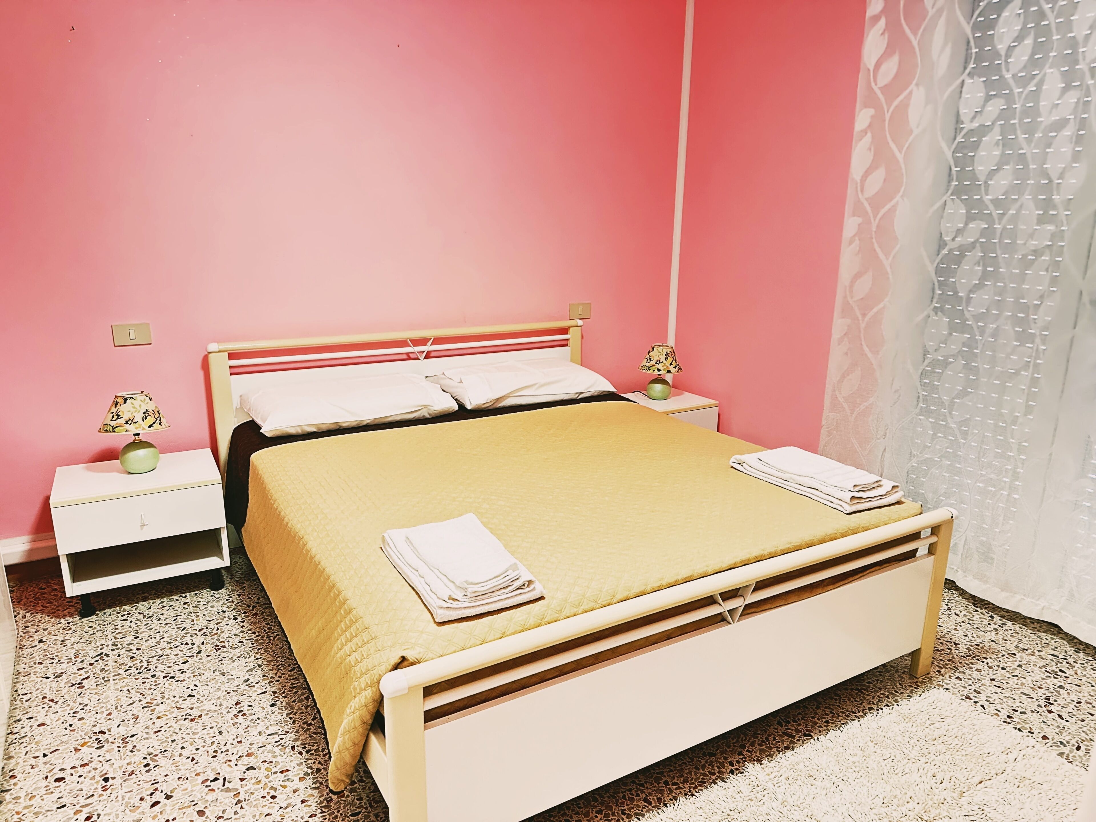 A single-bed room with a wooden frame, a pink wall, a patterned nightstand, and a white curtain.