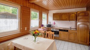 Een houten interieur met een eettafel, stoelen, een keukenhoek met kastjes, een magnetron en een raam met uitzicht op groen.