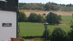 Een gebouw met een bord waarop 'Pension Wiebelhaus Mesler' staat, een bankje, een lantaarnpaal en uitzicht op een groen veld met bomen en een tractor in de verte.