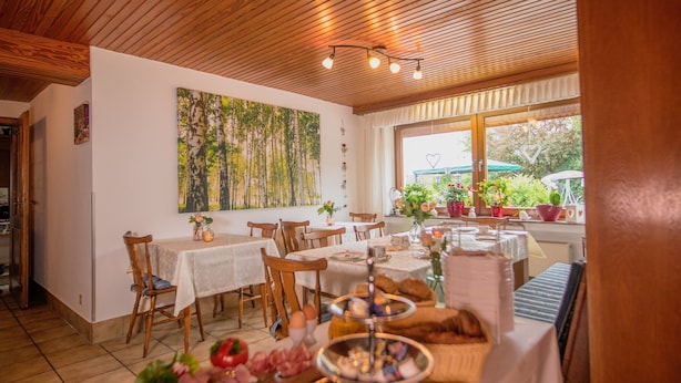 A dining area with wooden tables and chairs, a large painting on the wall, and a view through the window.