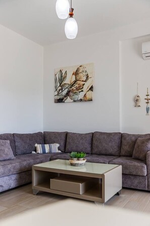 Een moderne woonkamer met een grijze modulaire bank, een glazen salontafel met planten, een aan de muur hangend schilderij en een plafondlamp.