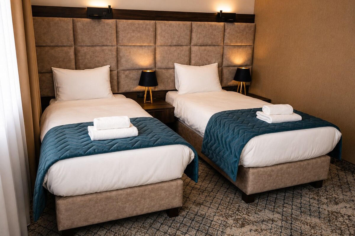 Habitación de hotel con dos camas, cada una con almohada, una mesita de noche con lámpara y una manta acolchada.