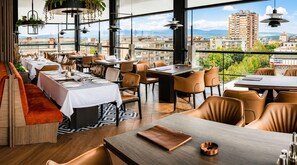 A restaurant with multiple tables and chairs, a wooden bench, and large windows offering a city view. - Hotel Leskovac (Leskovac)