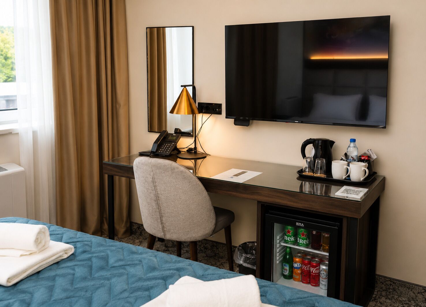 Ett hotellrum med en säng, skrivbord, stol, TV, lampa, telefon, kaffebryggare och minibar.