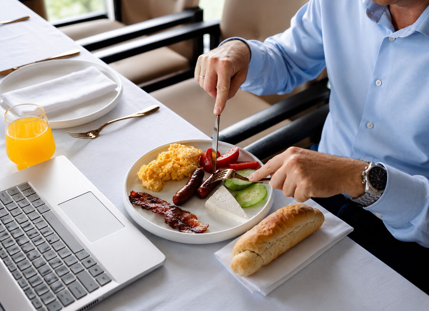 A breakfast setting with a laptop, a plate of food including eggs, bacon, tomatoes, and a baguette, and a glass of orange juice.