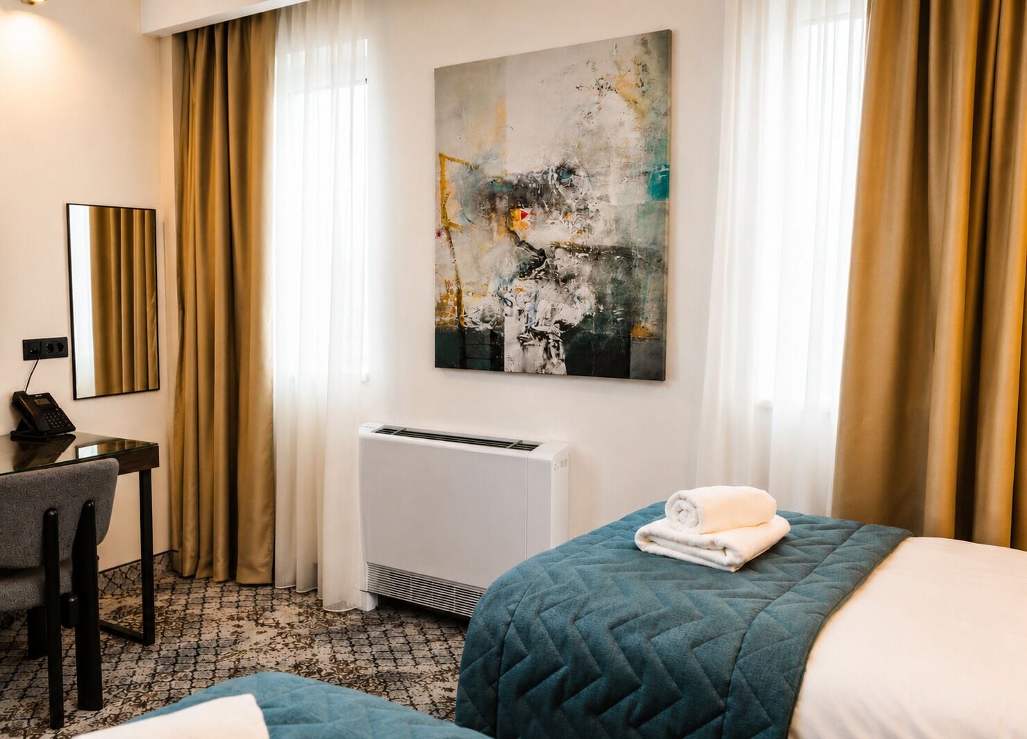 A hotel room with a bed, a desk with a telephone, a radiator, and a large abstract painting on the wall.