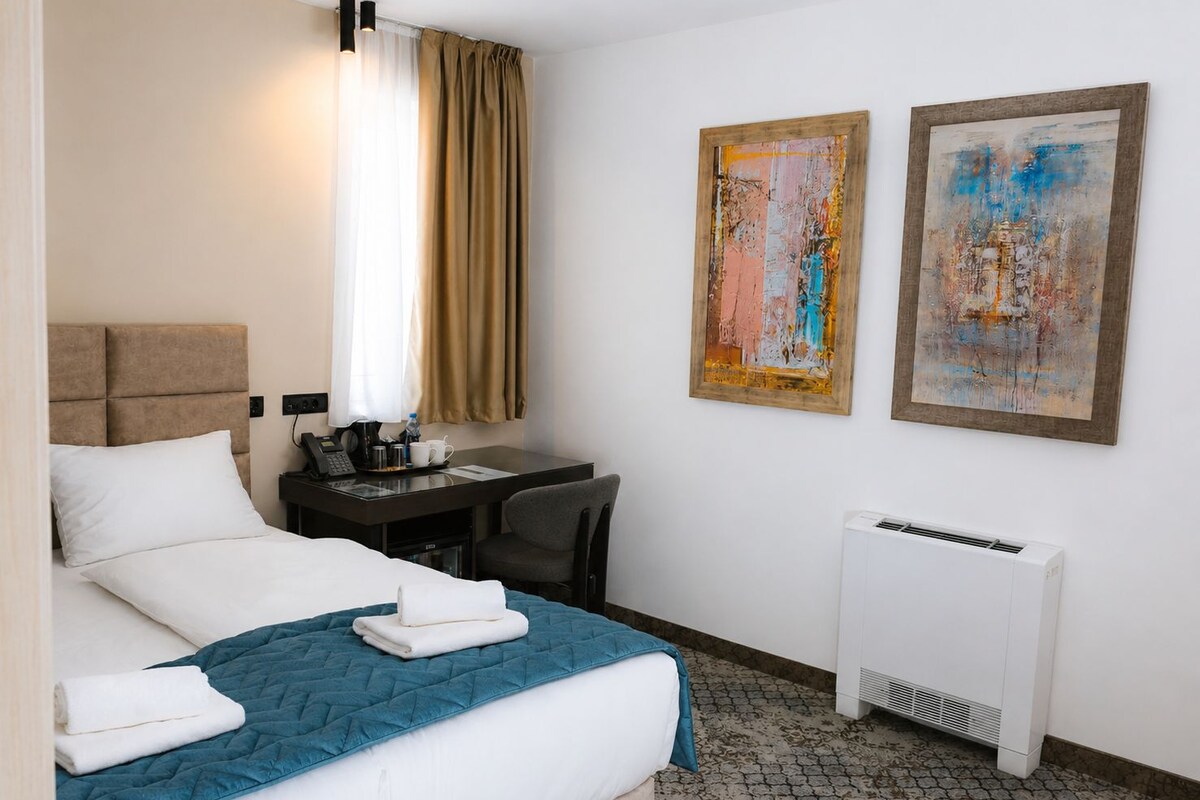 Una habitación de hotel con cama, escritorio, silla, dos cuadros y un aire acondicionado.